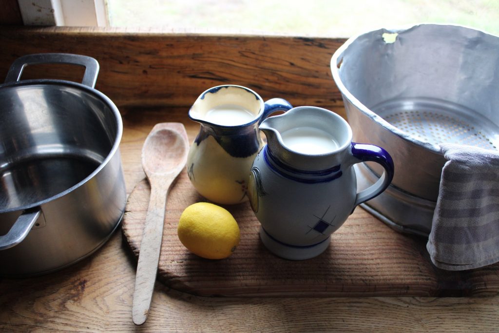 two jugs of milk, a lemon, a wooden spoon, a pot and a sieve with a dish towel - ingredients for how to make cream cheesem