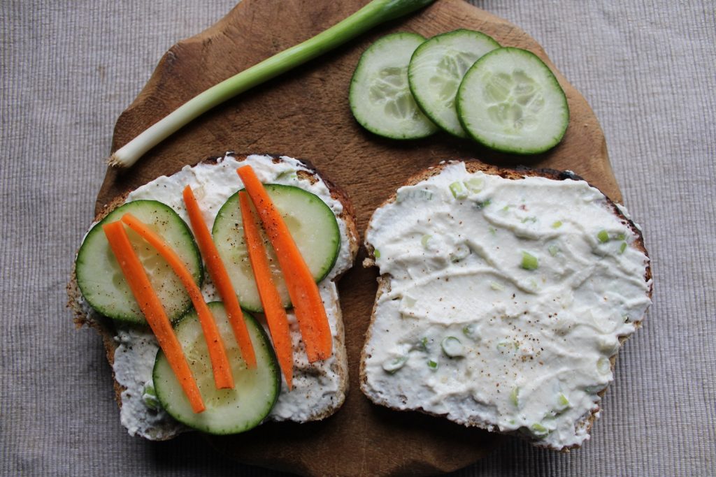 two slices of bread with homemade cream cheese, cucumber slices, carrot sticks and spring onion on a wooden board - how to make cream cheese