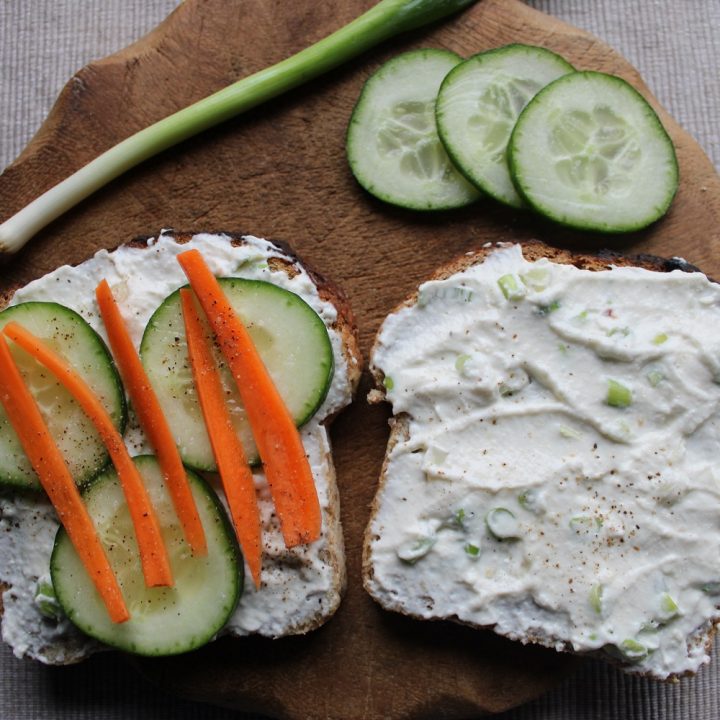 two slices of bread with homemade cream cheese, cucumber slices, carrot sticks and spring onion on a wooden board - how to make cream cheese